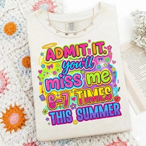 Peut inclure: T-shirt crème avec le texte "ADMIT IT. you'll miss me 6-7 TIMES THIS SUMMER" aux couleurs vives. Le t-shirt est orné d'un collier à petits charms. Le t-shirt est présenté sur une couverture crochetée.