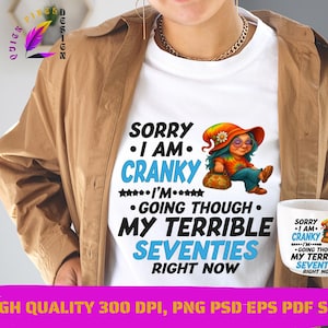 Peut inclure: T-shirt blanc avec le texte "Sorry I am cranky I'm going through my terrible seventies right now" et une illustration de dessin animé. Une tasse assortie est tenue à la main. L'image comprend également une veste marron.