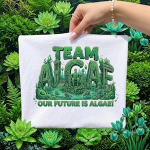 May include: A white t-shirt featuring a green graphic print with the text "TEAM ALGAE" and "OUR FUTURE IS ALGAE!" The design depicts a figure sitting on a throne surrounded by architectural structures and aquatic elements in various shades of green.