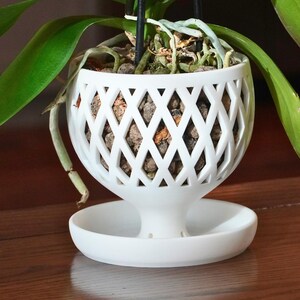 May include: White orchid planter with a diamond-shaped lattice design. The planter has a saucer base and is filled with brown orchid bark.