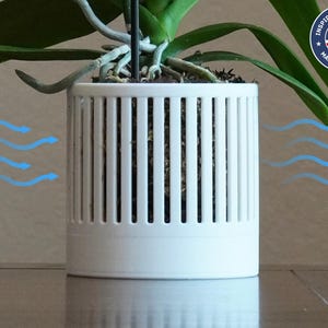 Puede incluir: Una maceta cilíndrica blanca para orquídeas con ranuras verticales, que contiene una planta de orquídea. La maceta está diseñada pensando en el flujo de aire, como indican las flechas azules. Fabricado en EE. UU., como indica el logotipo.