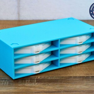 Puede incluir: Una caja de almacenamiento de plástico azul brillante con ocho compartimentos. Cada compartimento tiene una bandeja extraíble blanca. La caja está diseñada para guardar objetos pequeños como joyas, suministros de manualidades u otros objetos pequeños.