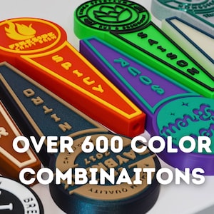 May include: A close-up of colorful, 3D printed beer tap handles with various designs and text, including "Fireside Craftworks", "Nitro", "Sails", "Across", "Mango Dreams", and "Lager". The text "OVER 600 COLOR COMBINATIONS" is displayed below the handles.