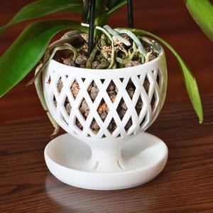 May include: White ceramic orchid pot with a lattice design and a matching saucer. The pot has a wide opening and a narrow base, and the saucer is round and flat. The pot is filled with brown orchid bark and the roots of an orchid plant are visible.