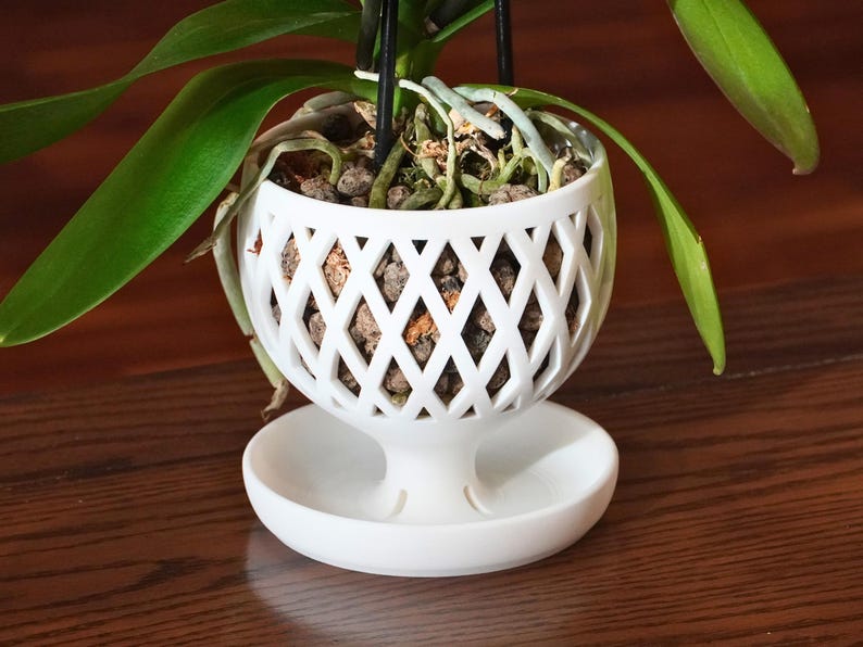 May include: White ceramic orchid pot with a lattice design and a matching saucer. The pot has a wide opening and a narrow base, and the saucer is round and flat. The pot is filled with brown orchid bark and the roots of an orchid plant are visible.