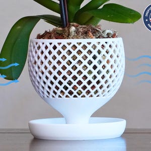 May include: White orchid plant in a white, open-weave, round pot with a white saucer. The pot has a unique design with a diamond pattern. The plant is sitting on a wooden surface. The image shows air flowing around the plant.