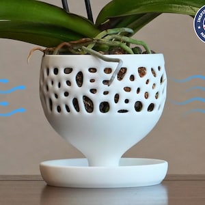 May include: White ceramic orchid planter with a decorative, openwork design. The planter has a wide base and a narrow opening at the top. The planter is designed to allow air to circulate around the roots of the orchid.