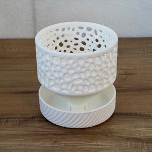 May include: White ceramic planter with a decorative, openwork design. The planter has a lower section with a textured, ribbed design.