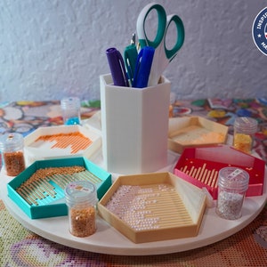 Puede incluir: Una bandeja giratoria blanca con seis compartimentos para organizar los suministros de pintura de diamantes. Cada compartimento tiene una bandeja pequeña con una cuadrícula de agujeros para contener las cuentas. También hay varios frascos pequeños llenos de cuentas y un portaplumas con bolígrafos y tijeras.