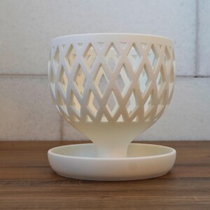 May include: White ceramic planter with a diamond-shaped lattice design. The planter has a wide, shallow base and a narrow, cylindrical top.