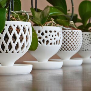May include: Four white ceramic orchid pots with different patterned designs. The pots are hanging from the ceiling and have a saucer underneath.