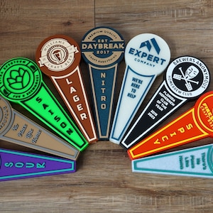 May include: A collection of 10 colorful tap handles with various brewery names and logos. The tap handles are made of wood, metal, and plastic. The tap handles are arranged in a semi-circle on a wooden surface.