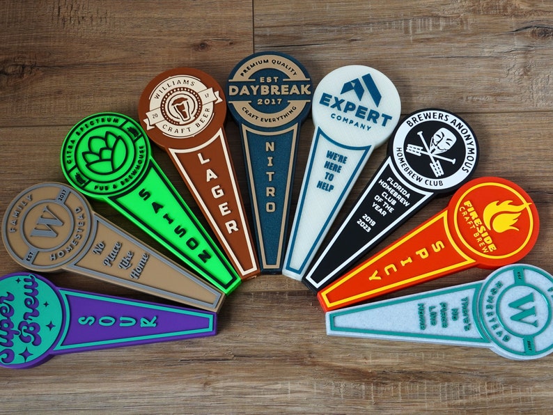 May include: A collection of 10 colorful tap handles with various brewery names and logos. The tap handles are made of wood, metal, and plastic. The tap handles are arranged in a semi-circle on a wooden surface.