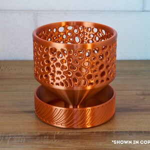May include: A copper-colored planter with a geometric pattern and a textured base. The planter is designed for indoor plants and has a drainage system.