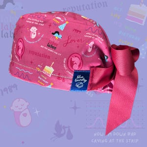 K&ouml;nnte beinhalten: Eine rosafarbene chirurgische M&uuml;tze mit einem bunten Muster, das ein Baby, einen Kuchen und den Text "labor labor labor", "Lover", "reputation", "1989", "Act Like It's Your Birthday Every Day", "Now I'm Down Bad Crying at the Strip" und "Head First Fearless" zeigt. Die M&uuml;tze hat ein blaues Etikett mit dem Text "blue laundry".