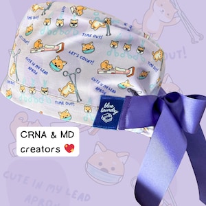 Può includere: Un berretto chirurgico bianco e viola con un simpatico motivo di cane Shiba Inu. Il berretto ha un nastro viola legato intorno e un'etichetta blu che dice "blue laundry". Il berretto ha il testo "Cute in my lead apron" e "Time out!" ripetuto su tutto il motivo.