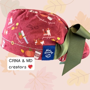 May include: A burgundy scrub cap with a green bow. The cap features a pattern of puppies, pumpkins, coffee cups, and medical-themed illustrations. The text "pumpkin spice" and "blue laundry" are also visible.