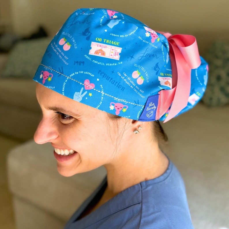 May include: Blue surgical scrub hat with a pink tie. The hat has a variety of colorful patterns and text, including "OB Triage", "Lover", "reputation", and "Magic, Madness, Heaven, Sir".