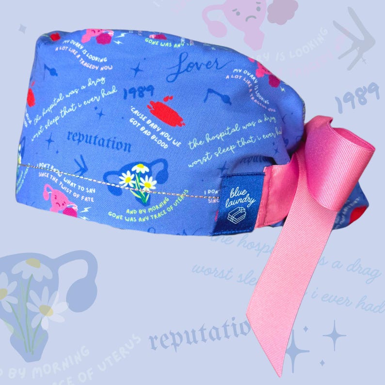 K&ouml;nnte beinhalten: Eine blaue OP-M&uuml;tze mit einer rosa Schleife. Die M&uuml;tze hat ein wei&szlig;es und rosa Blumenmuster und Text, der "Lover", "1989", "reputation", "the hospital was a drag", "worst sleep that I ever had", "cause baby now we got bad blood", "my ovary is looking a lot like a tragedy now", "gone was any trace of uterus", "I don't sing", "blue laundry" lautet.