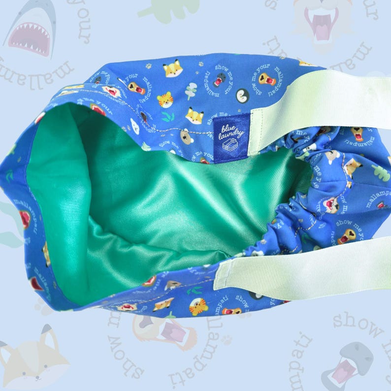 Puede incluir: Un gorro de tela azul con interior verde azulado y correas el&aacute;sticas blancas. El exterior presenta un estampado de zorros y perros de dibujos animados, con el texto "show me your mallampati". Una peque&ntilde;a etiqueta azul dice "blue laundry".