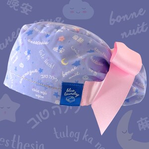 May include: A light purple scrub cap with a pink bow. The cap is patterned with stars, moons, and text in multiple languages. A blue tag reads "blue laundry".