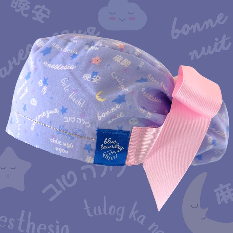 May include: A light purple scrub cap with a pink bow. The cap is patterned with stars, moons, and text in multiple languages. A blue tag reads "blue laundry".