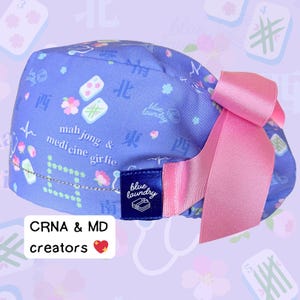 Peut inclure: Un bonnet de chirurgien bleu clair avec un nœud rose. Le bonnet présente un motif de tuiles de Mahjong, de fleurs et de caractères chinois. Le texte "mahjong & medicine girlie" est imprimé sur le bonnet, ainsi que le logo "blue laundry".