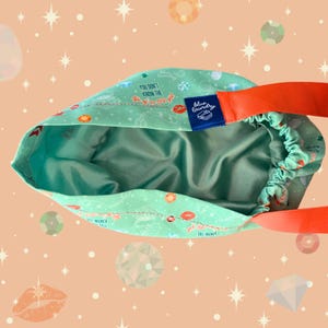 May include: A turquoise drawstring bag with an orange strap. The bag's exterior features a pattern of lips, donuts, and text that reads "You Don't Know The F*ck Or Girl". The interior is a solid teal color, and a blue label reads "Blue Laundry".
