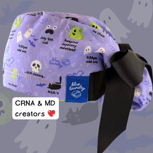 May include: A light purple scrub cap with a black bow. The cap features a repeating pattern of ghosts, bats, and humorous phrases like "hospital mystery meatloaf." A blue tag reads "blue laundry." Text on the cap includes "CRNA & MD creators."