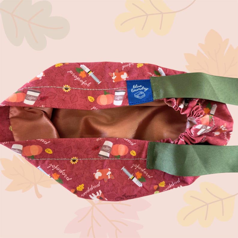 May include: A burgundy tote bag with a repeating pattern of pumpkins, coffee cups, syringes, and corgis. The bag has a green strap and a blue label that says "blue laundry". The interior is a copper color.