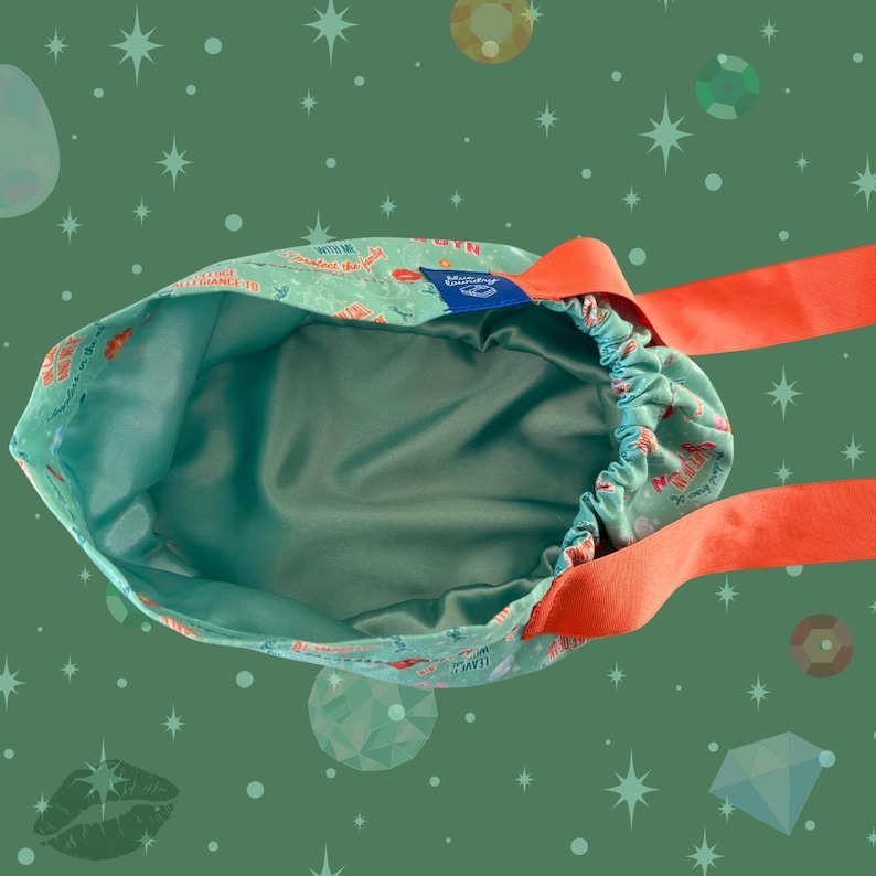 May include: A teal drawstring bag with a satin interior and orange ribbon straps. The exterior features a repeating pattern of red and white text and illustrations. A blue tag is sewn on the side.