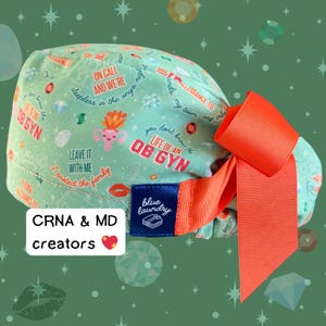 May include: A light green scrub cap with an orange bow. The cap features text and graphics related to obstetrics and gynecology, including the words "OB GYN" and "Life of an OB GYN". The cap also has the text "blue laundry" on a blue tag.