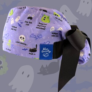 May include: A lavender scrub cap with a black bow. The cap is patterned with cartoon ghosts, bats, and text including "July 4th call" and "hospital mystery meatloaf." A blue label reads "blue laundry."