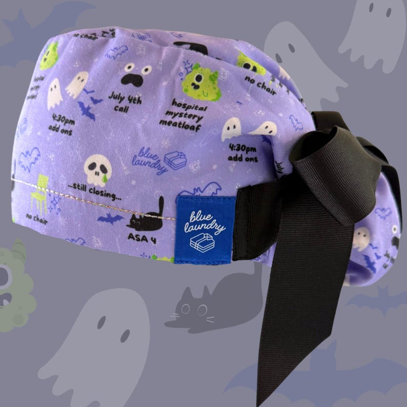 May include: A lavender scrub cap with a black bow. The cap is patterned with cartoon ghosts, bats, and text including "July 4th call" and "hospital mystery meatloaf." A blue label reads "blue laundry."