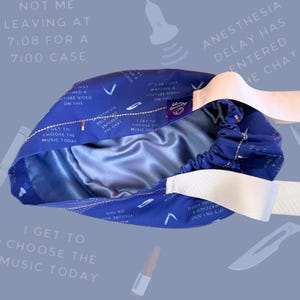May include: A blue surgical cap with white elastic straps. The cap has a satin lining and features a repeating pattern of text and images, including "I get to choose the music today" and "Anesthesia delay has entered the chat".