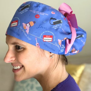 May include: A blue scrub cap with a pink bow, featuring various illustrations and text. The cap includes the text "HELLO MY NAME IS Anesthesia", "1989", and other phrases. The design incorporates images of a guitar, lipstick, and a remote control.