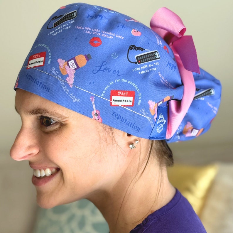 May include: A blue scrub cap with a pink bow, featuring various illustrations and text. The cap includes the text "HELLO MY NAME IS Anesthesia", "1989", and other phrases. The design incorporates images of a guitar, lipstick, and a remote control.