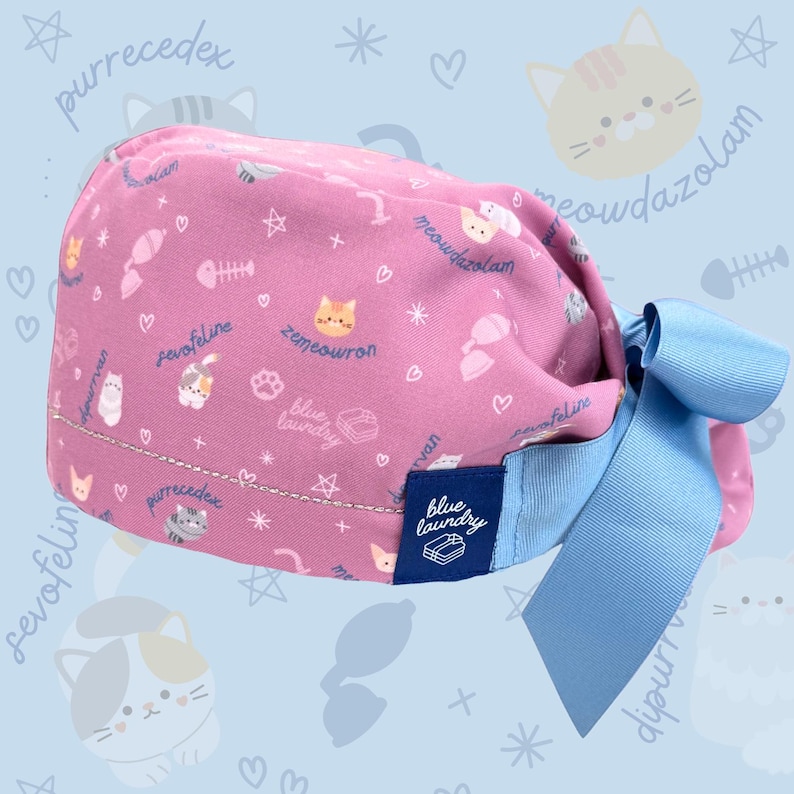 May include: A pink scrub cap with a light blue bow. The cap is patterned with cat illustrations, fish bones, and text. A blue label with the text "blue laundry" is sewn on the side. The cap is designed for medical professionals.