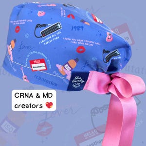 Puede incluir: Un gorro quir&uacute;rgico azul con una cinta rosa. El gorro presenta varios gr&aacute;ficos, incluyendo un mando a distancia, labios y una guitarra. El texto incluye "Lover", "Anesthesia" y "1989".