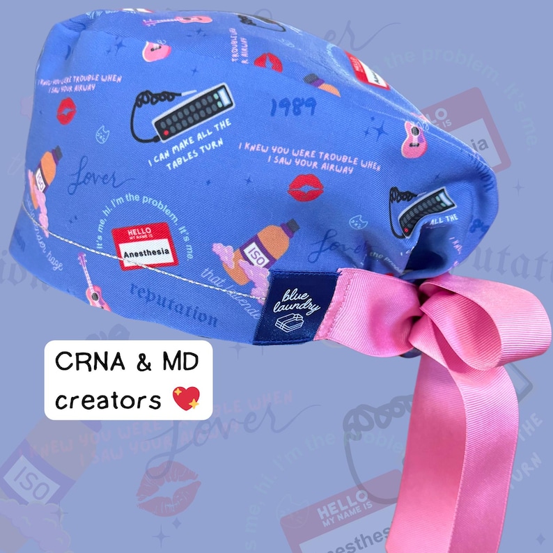 Puede incluir: Un gorro quir&uacute;rgico azul con una cinta rosa. El gorro presenta varios gr&aacute;ficos, incluyendo un mando a distancia, labios y una guitarra. El texto incluye "Lover", "Anesthesia" y "1989".