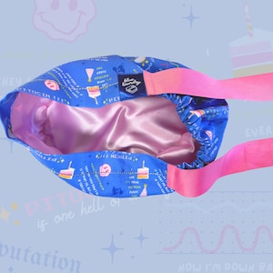 K&ouml;nnte beinhalten: Ein blaues Satin-Haargummi mit einem rosa Band. Das Haargummi hat ein Geburtstagstortenmuster mit dem Text "PITOCIN!!" und "Es ist jeden Tag dein Geburtstag!" darauf gedruckt.