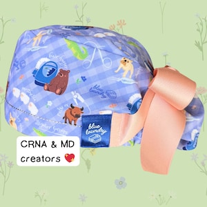 May include: A blue and white patterned scrub cap with a peach-colored bow. The cap features illustrations of animals and medical tools. The text "CRNA & MD creators" is visible on the image.