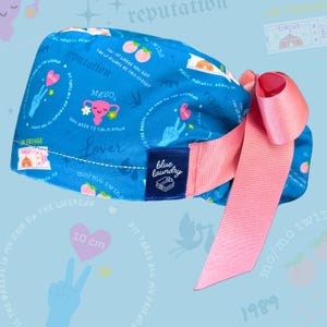 K&ouml;nnte beinhalten: Eine blaue OP-M&uuml;tze mit einer rosa Schleife und verschiedenen bunten Mustern, darunter Pfirsiche, Herzen, Peace-Zeichen und der Text "reputation", "Lover", "mo/mo twins", "1989", "Magic Madness", "Heavier Than Heaven", "Can I Go Where You Got", "Can We Always Be This Close?", "You Need To Calm Down", "Pit Takes All My Friends", "It's The Breeze In My Hair On The Weekend", "10 cm" und "blue laundry".