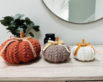 May include: Three knitted pumpkins in shades of orange, gray, and white, each with a cinnamon stick stem and a velvet ribbon bow. The pumpkins are arranged on a wooden surface, suggesting a fall or harvest theme.