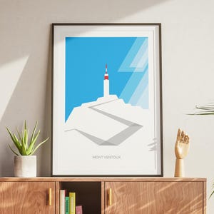 May include: Framed art print depicting a minimalist illustration of Mont Ventoux, with a white mountain and a blue sky. The artwork includes a white tower with a red top and the text "MONT VENTOUX" at the bottom. The print is displayed on a wooden cabinet.
