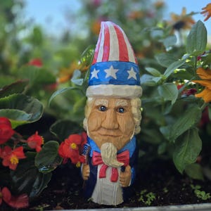 Könnte beinhalten: Eine geschnitzte Holzfigur von Uncle Sam mit einem rot-weiß-blauen Hut mit Sternen, einem blauen Mantel und einer roten Fliege. Die Figur hält eine kleine amerikanische Flagge. Der Hintergrund ist verschwommen mit grünem Laub und roten Blumen.