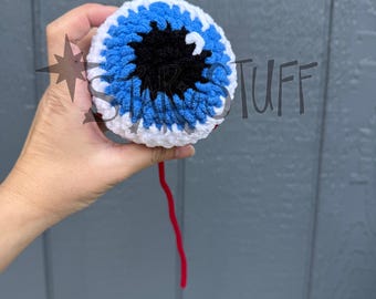 May include: A round, crocheted eyeball plush featuring a white outer ring, a bright blue iris, and a large black pupil with a small white highlight. A single thin red yarn strand hangs from the bottom. The text STAR STUFF is overlaid.