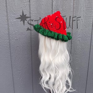 May include: A red and green crocheted hat with white bead accents and the text "STAR STUFF". The hat has a ruffled green brim and is displayed on a white wig. The hat is likely for costume or cosplay.