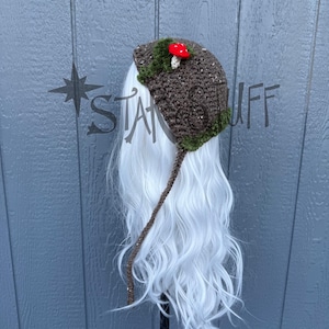 May include: A brown crochet hat with a red mushroom and green trim. The hat has a long, brown crocheted strap. The hat is on a white wig. The hat is a whimsical accessory.