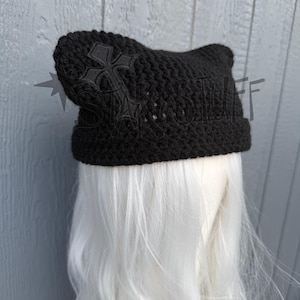 May include: A black crocheted beanie hat with cat ears. The hat features a cross design on the side. The hat is displayed on a white wig head against a gray background.